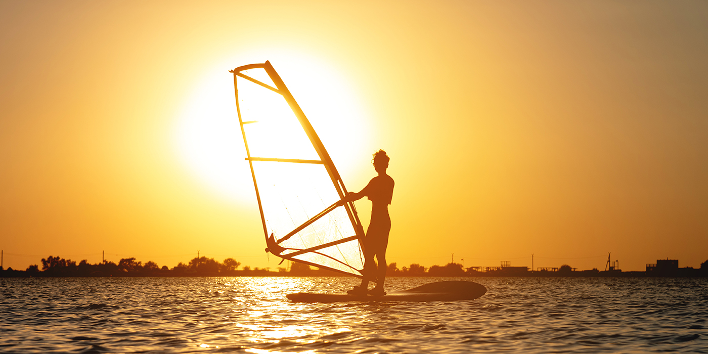 A beginner windsurfer woman stands on a board with a sail on a sunset background. Windsurfing school